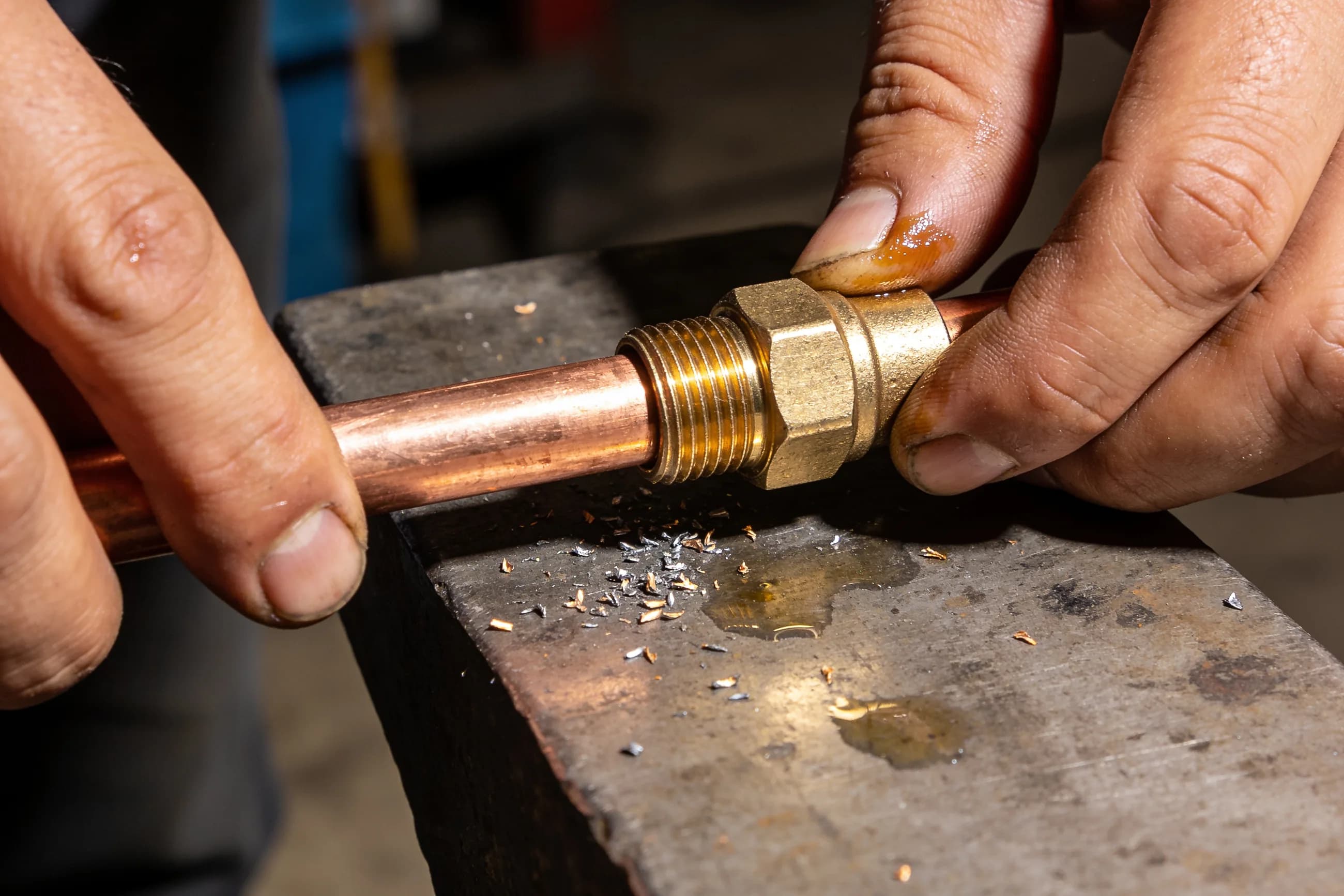 A machinist threading a solid brass compression fitting onto a length of copper pipe over a steel bench.